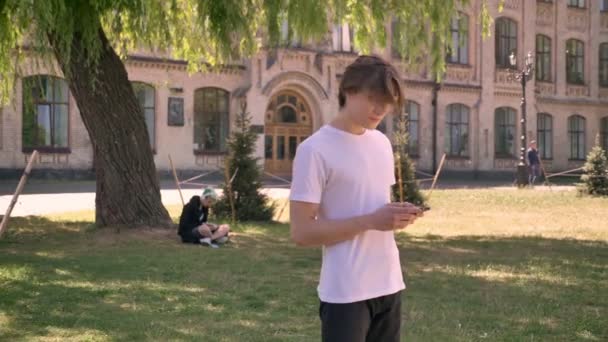 Молодий гарненький студент-чоловік у білій сорочці стоїть і тримає телефон, парк біля університету фону — стокове відео