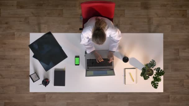 Jeune scientifique en blouse blanche tapant sur ordinateur portable et au téléphone avec chromatique, topshot, assise à table avec balayage par rayons X, concept médical — Video