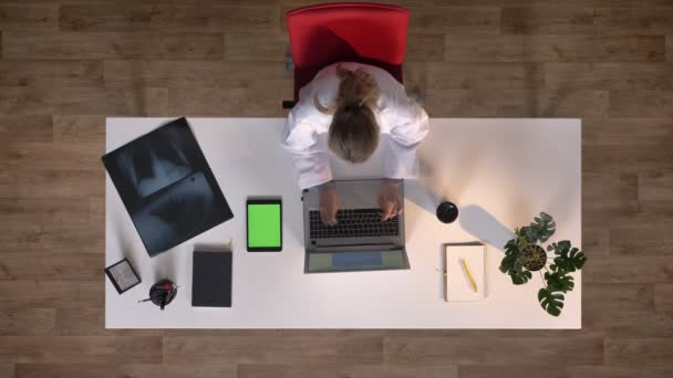 Enfermera joven escribiendo en el ordenador portátil y en la tableta con chromakey, sentado a la mesa con rayos X escaneo, concepto médico, topshot — Vídeo de stock