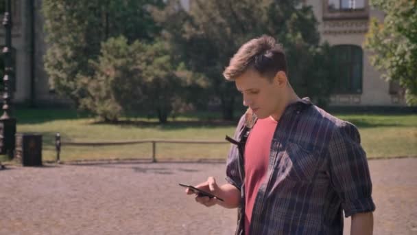 Joven hombre guapo está caminando por el parque durante el día en verano, tocando en el teléfono inteligente, concepto de comunicación — Vídeo de stock