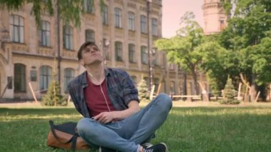 Genç yakışıklı adam parkta çimlerin üzerine oturuyor, kulaklık ile müzik dinleme, kavram, kavram, arka planda bina rüya sakin ol