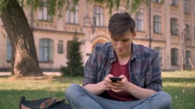 Genç yakışıklı adam parkta çimlerin üzerine oturuyor, smartphone dokunarak, kavram, iletişim kavramı, arka planda bina sakin ol