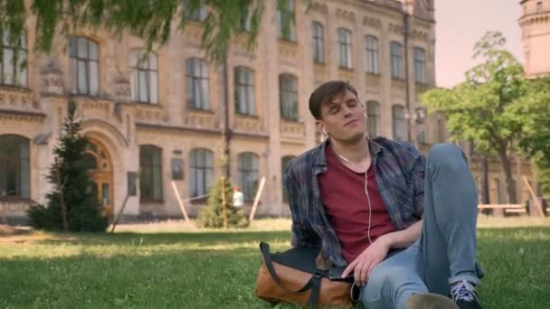 若いハンサムな男が公園の芝生の上に座っている、イヤホンで音楽を聞いて、リラックス背景に建物のコンセプト — ストック動画