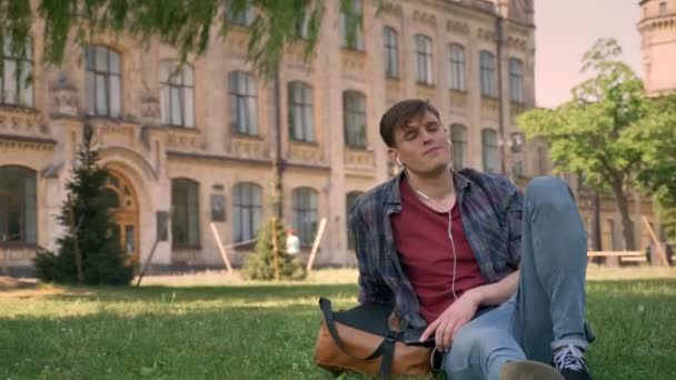 Joven hombre guapo está sentado en la hierba en el parque, escuchando música con auriculares, relajarse concepto, la construcción de fondo — Vídeo de stock