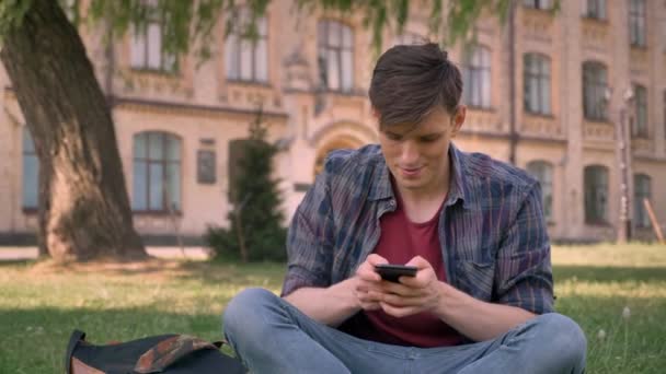 Όμορφος νεαρός άνδρας κάθεται στο γρασίδι στο πάρκο, πατώντας στο smartphone, χαλαρώστε έννοια, έννοια επικοινωνίας, κτίριο σε φόντο — Αρχείο Βίντεο