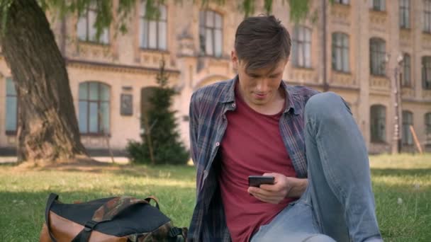 Genç yakışıklı adam parkta çimlerin üzerine oturmuş, Internet sayfalarında Smartphone cep telefonu ile izlerken, kameraya izlerken, kavram, iletişim kavramı, arka planda bina sakin ol — Stok video