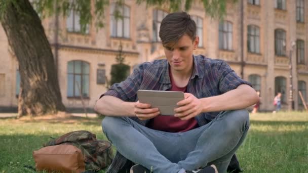 Joven hombre guapo está sentado en la hierba en el parque, viendo páginas en Internet en la tableta, mirando a la cámara, concepto de relajación, concepto de comunicación, edificio en el fondo — Vídeo de stock