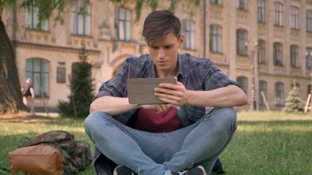 El joven está sentado en la hierba en el parque, viendo páginas en Internet en la tableta, mirando a la cámara, concepto de relajación, concepto de comunicación, edificio en el fondo — Vídeo de stock