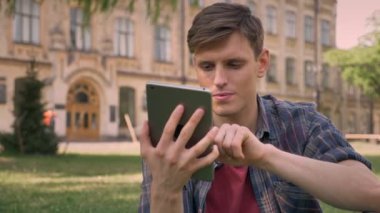 Genç yakışıklı adam parkta çimlerin üzerine oturuyor, tablet, musluklar mavi ekran gösterir kameraya izlerken, kavram, iletişim kavramı, arka planda bina sakin ol