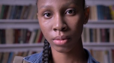 Genç Afrikalı Amerikalı kız öğrenci kameraya bakıyor ve kütüphanede duruyor, ciddi ve endişeli.