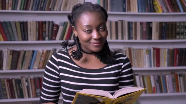 Jeune belle femme afro-américaine lisant un livre à la bibliothèque, souriante, heureuse, fond de bibliothèque — Video