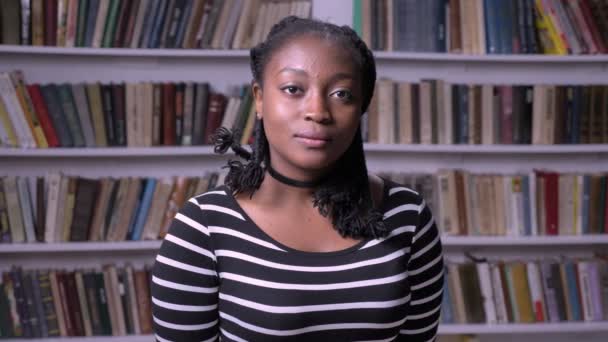 Красивий афро-американських брюнетки стоячи в бібліотеці і дивлячись на камеру, серйозні і замислений, книжкові полиці фону — стокове відео