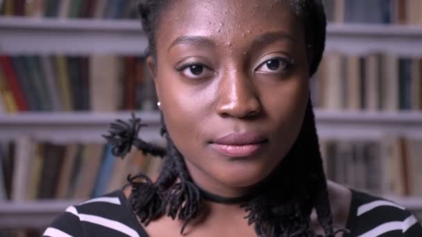 Mujer afroamericana joven mirando a la cámara y de pie en la biblioteca, preocupado y serio, librerías fondo — Vídeos de Stock