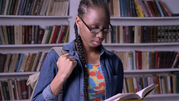 Jovem mulher americana africana bonita em óculos livro de leitura na biblioteca e olhando para a câmera, feliz, sorrindo, prateleiras de fundo — Vídeo de Stock