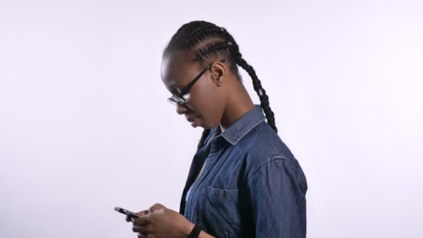 Young african american woman in glasses texting on phone and looking at camera, isolated over white background, smiling, happy — Stock Video