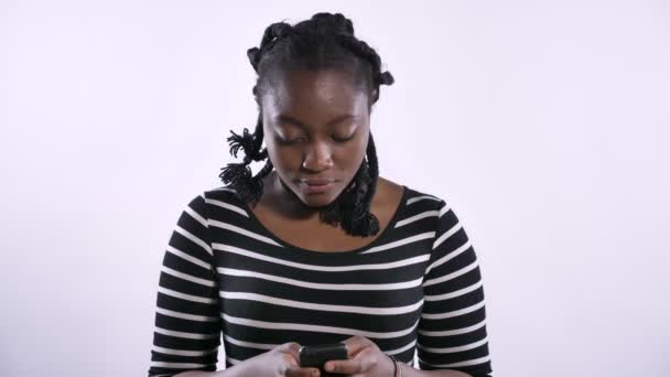 Jonge mooie Afrikaanse Amerikaanse vrouw te typen op de telefoon, camera kijken en lachend, blij, witte studio achtergrond — Stockvideo