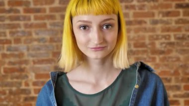 Kameraya bakarak ve tuğla arka plan, gülümseyen ve pozitif duran sarı saçlı Genç büyüleyici kadın