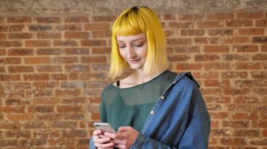 Telefonda yazarak ve kamera, tuğla arka plan, mutlu ve neşeli ayakta gülümseyen sarı saç modeli olan güzel kadın
