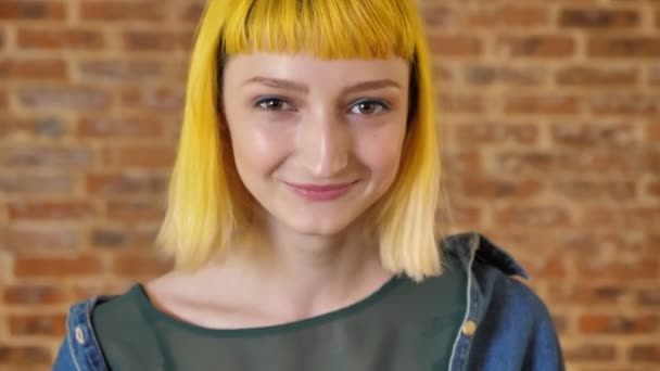 Portrait de jeune belle femme aux cheveux jaunes regardant la caméra et souriant, fond de mur de briques, heureux et joyeux — Video