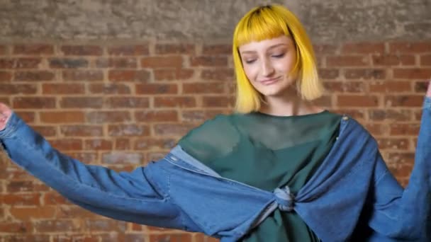 Joven mujer inusual con el pelo amarillo bailando y sonriendo a la cámara, aislado sobre el fondo de ladrillo, feliz — Vídeos de Stock