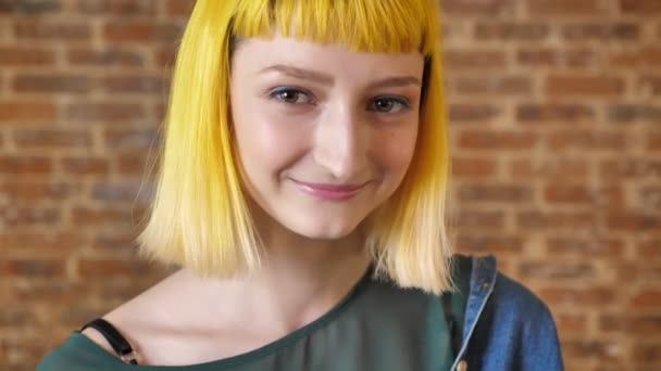 Молодая застенчивая женщина с желтыми волосами смотрит в камеру и улыбается, кирпичный фон стены, веселый и счастливый — стоковое видео