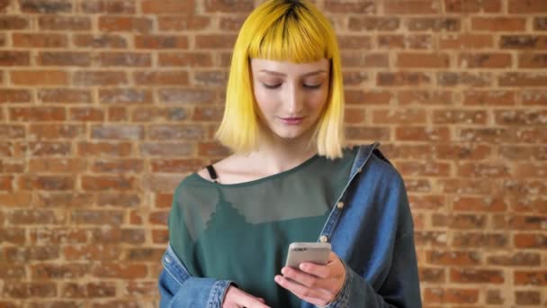 Giovane donna affascinante con i capelli gialli digitando sul telefono e sorridente, in piedi vicino al muro di mattoni sfondo, persona insolita, straordinario — Video Stock