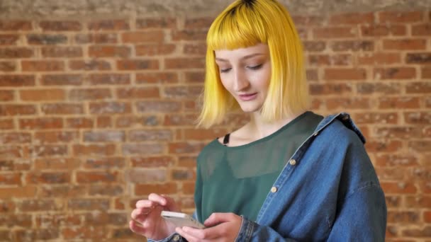 Jeune femme inhabituelle avec des cheveux jaunes tenant le téléphone et regardant la caméra, souriant et debout près du fond du mur de briques — Video