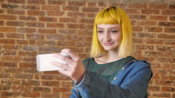Ung charmig kvinna med gult hår tar selfie med telefon, leende och står nära tegel vägg bakgrund, modern ungdom — Stockvideo