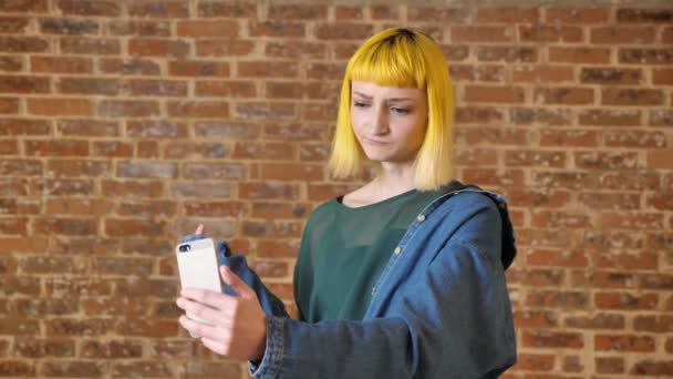 Jonge vrouw met gele haren praten met iemand via video chat, staande in de buurt van baksteen achtergrond — Stockvideo