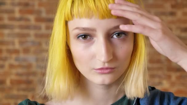 Portrait de jeune femme aux cheveux jaunes regardant la caméra et bougeant ses mains autour de son visage, fond de mur de briques — Video