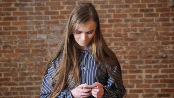 Menina bonita nova está assistindo seu smartphone, conceito de comunicação, conceito relaxante, fundo de tijolo — Vídeo de Stock