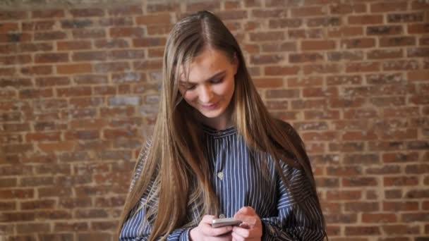Menina bonita nova está assistindo seu smartphone, sorrindo, conceito de comunicação, conceito relaxante, fundo de tijolo — Vídeo de Stock
