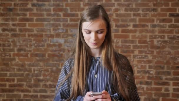 Giovane bella ragazza sta guardando il suo smartphone, concetto di pensiero, concetto di comunicazione, concetto di relax, sfondo mattone — Video Stock