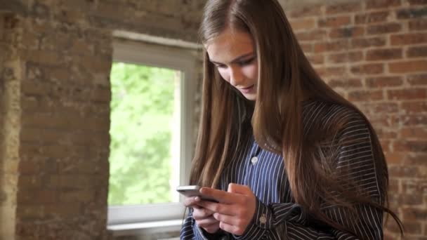 Ung vacker flicka tittar på sin smartphone, kommunikationskoncept, tegel bakgrund — Stockvideo