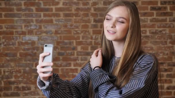 Joven hermosa chica está haciendo selfie en su teléfono inteligente, sonriendo, tocando el cabello, concepto de comunicación, fondo de ladrillo — Vídeos de Stock