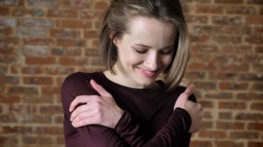 Genç Güzel utangaç kız kameraya, izliyor gülümseyerek, aşağı, izlerken kendini tuğla arka plan hugs
