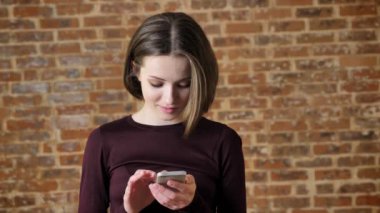 Genç neşeli kız kameraya, gülümseyen, iletişim kavramı, tuğla arka plan izlerken onun smartphone, ileti yazmakta olduğunu