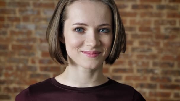 Young beautiful confident girl is watching at camera, smiling, brick background — Stock Video