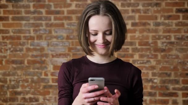 Menina bonita jovem está lendo mensagem em seu smartphone, sorrindo, conceito de comunicação, fundo de tijolo — Vídeo de Stock