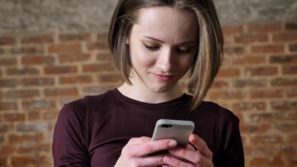 Joven hermosa chica está leyendo el mensaje en su teléfono inteligente, riendo, concepto de comunicación, fondo de ladrillo — Vídeos de Stock