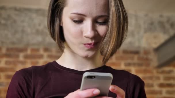 Joven hermosa chica está escribiendo mensaje en su teléfono inteligente, sonriendo, concepto de comunicación, fondo de ladrillo — Vídeo de stock