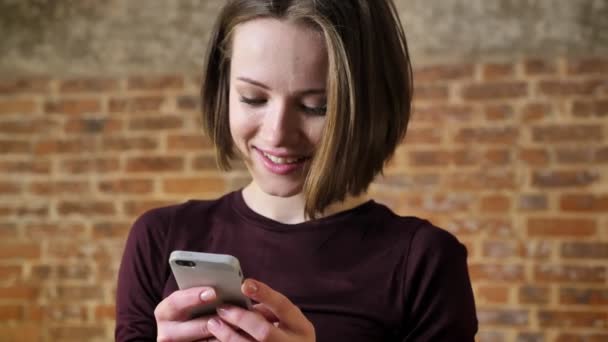 Joven linda chica está leyendo el mensaje en su teléfono inteligente, se sorprende, concepto de comunicación, fondo de ladrillo — Vídeo de stock