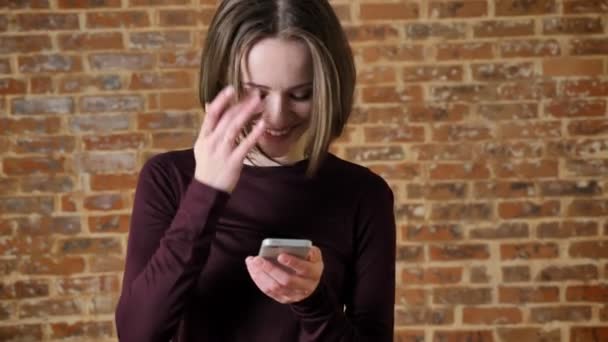 Giovane ragazza allegra sta digitando il messaggio sul suo smartphone, sorridente, concetto di comunicazione, sfondo di mattoni — Video Stock