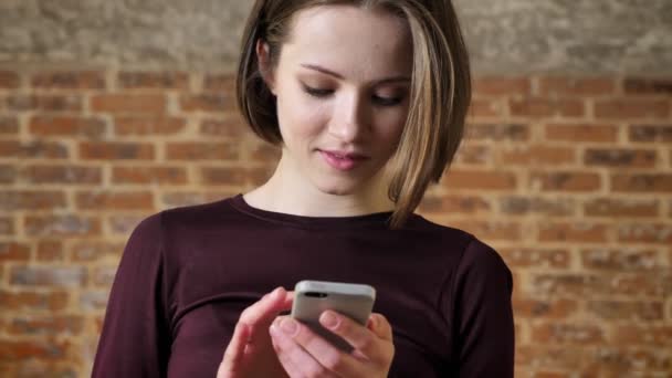 Joven chica atractiva está escribiendo mensaje en su teléfono inteligente, mirando a la cámara, sonriendo, concepto de comunicación, fondo de ladrillo — Vídeos de Stock