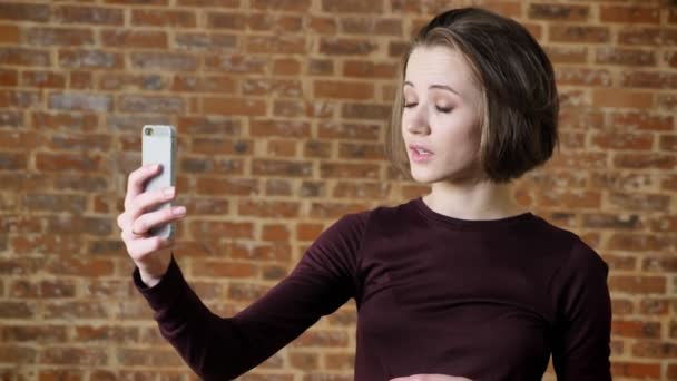 Menina bonita nova tem chamada de vídeo em seu smartphone, conceito de comunicação, fundo de tijolo — Vídeo de Stock