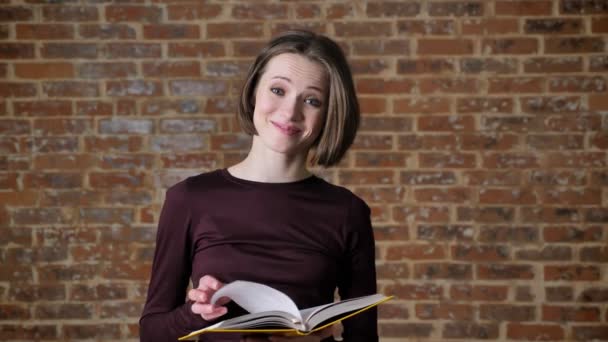 Menina agradável nova está lendo livro, assistindo a câmera, sorrindo, fundo de tijolo — Vídeo de Stock