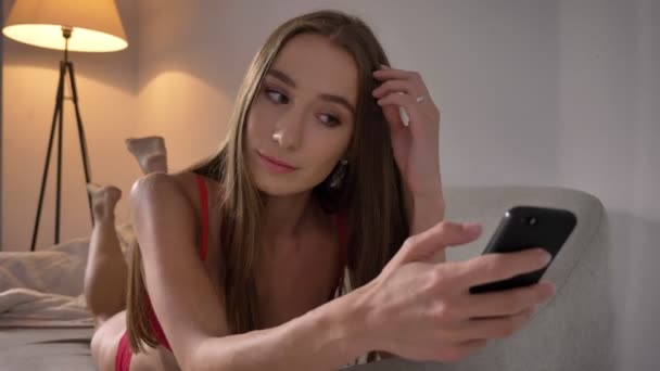 ソファやソファで、セクシーなかなり美しい女性の携帯電話にテキスト メッセージの上に横たわる下着姿の若い魅力的な女性 — ストック動画