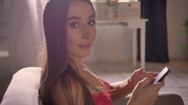 Genç kadın kamera başına dönme ve gülümseyen, smartphone holding, oturma odasında sandalyede oturan kırmızı sutyen