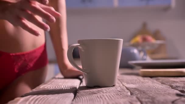 Close shot of womans hand taking cup from wooden table, perfecto cuerpo atractivo en el fondo, de pie en la cocina — Vídeo de stock