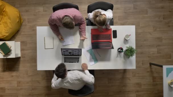 Tre giovani che lavorano sul computer portatile, un altro uomo che scrive sul documento in ufficio, topshot, seduto a tavola e digita sul computer — Video Stock